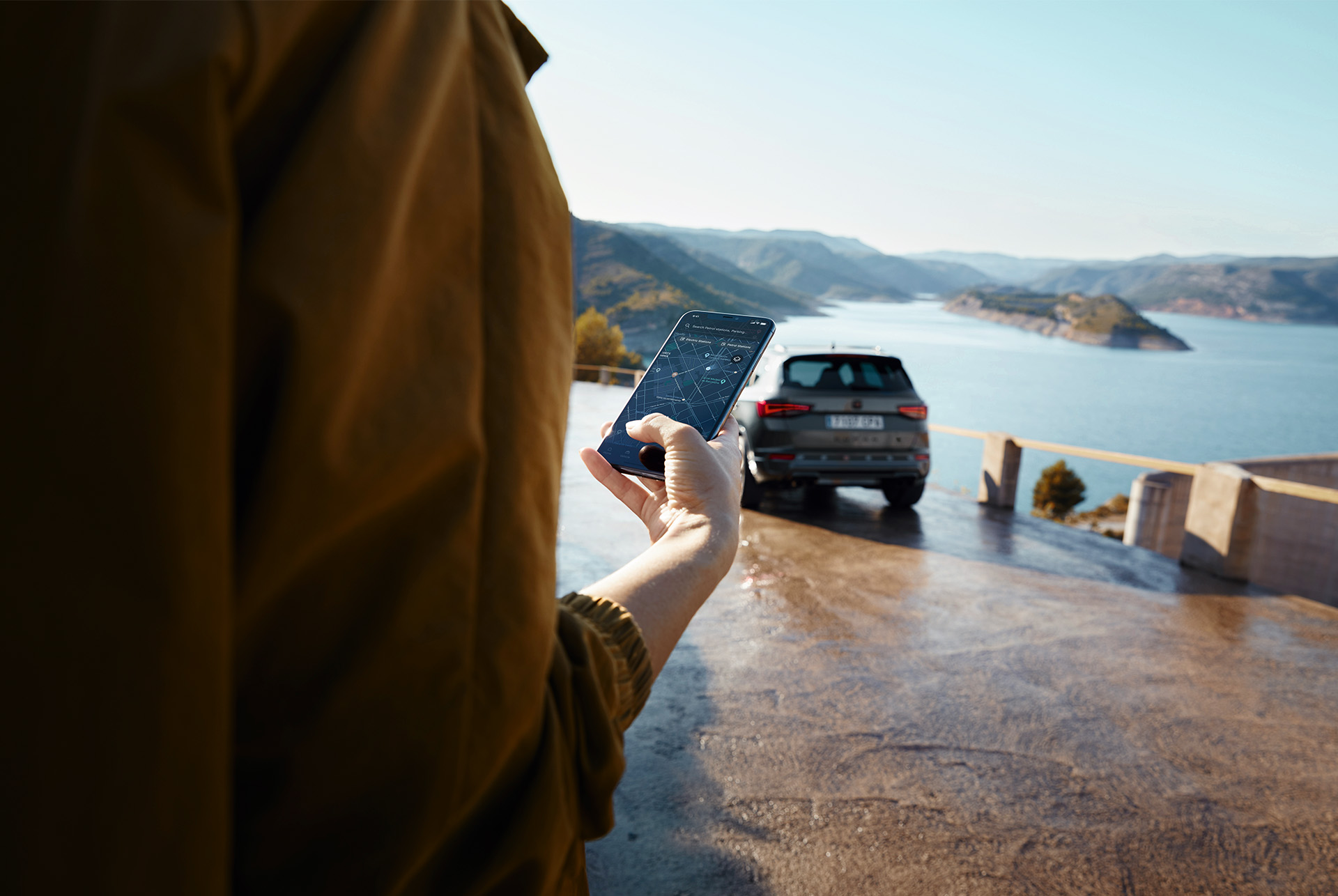 Une personne tenant un smartphone, la CUPRA Ateca en arrière-plan, une vue panoramique sur la montagne et un lac, symbolisant un monde connecté.