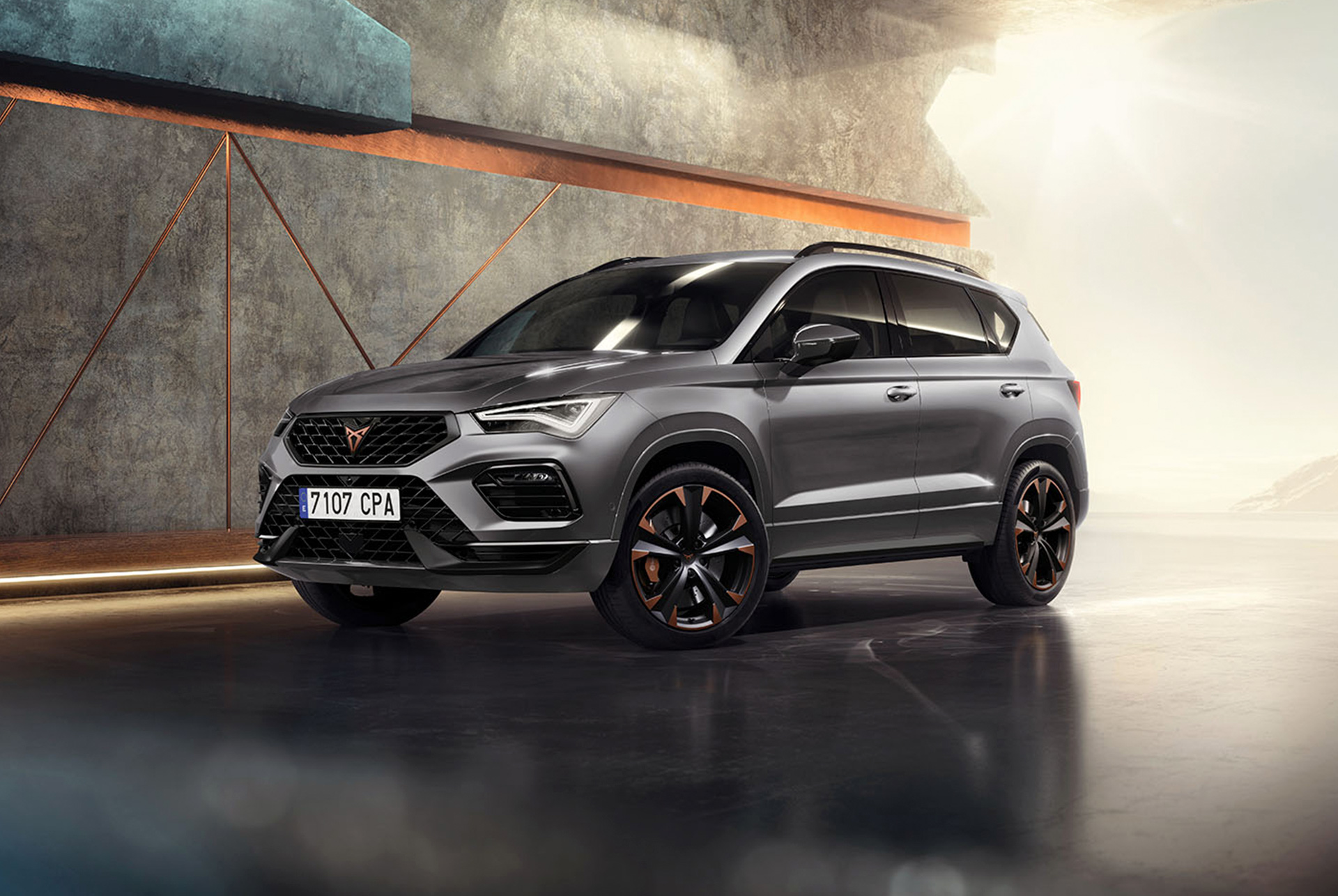 Cupra ateca 2024 en gris graphite, un SUV compact et sportif, garé dans un cadre moderne et industriel. Le design de la voiture se caractérise par des lignes aérodynamiques, des phares LED et des jantes en alliage distinctives aux accents cuivrés. L’arrière-plan se compose d’un mur géométrique qui contraste avec le design épuré du véhicule.